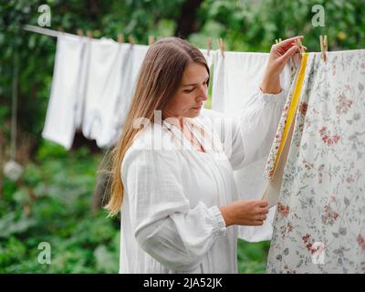 middle aged caucasian woman in white dress doing homework, hanging clothes on clothesline on street in courtyard of village cottage house, laundry day concept Stock Photo