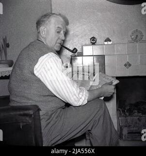 1950s, historical, early evening and an elderly gentleman sitting infront of a coal fire with a newspaper and smoking a pipe, England, UK. Stock Photo