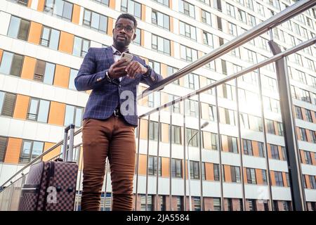 well dressed and successful African american businessman walking for flight at airport with wheeled bag suitcase looking at the phone screen Stock Photo