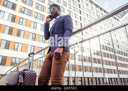 well dressed and successful African american businessman walking for flight at airport with wheeled bag suitcase looking at the phone screen Stock Photo