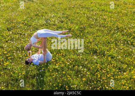 Beautiful young couple doing acro yoga in park. Man lying on grass and balancing woman in his feet. Stock Photo
