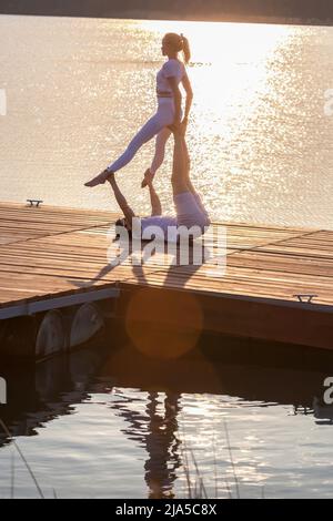 Beautiful young couple doing acro yoga on the pier against sunset. Man lying on the pier and balancing woman in his feet. Stock Photo