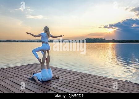 Beautiful young couple doing acro yoga on the pier against sunset. Man lying on the pier and balancing woman in his feet. Stock Photo