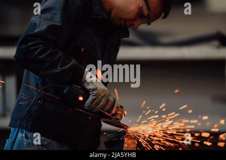 Heavy Industry Engineering Factory Interior with Industrial Worker Using Angle Grinder and Cutting a Metal Tube. Contractor in Safety Uniform and Hard Stock Photo