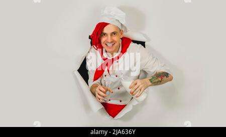 Man with red dreadlocks holds it in his hand milk. Confident young cook in white apron and red tie pours milk from a bottle into a glass, peeking thro Stock Photo