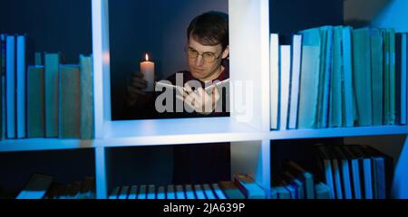 Man in glasses with candle reading book, standing behind bookshelf. Male holds burning candle while reading book in wooden bookcase with various liter Stock Photo