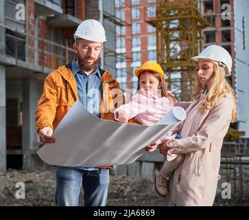Family with child studying architectural drawings outside apartment building under construction. Man and woman discussing building plan while standing with kid at construction site. Stock Photo