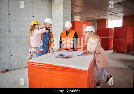 Family with child discussing architectural plan of new apartment with construction worker, standing inside apartment building under construction and talking with builder. Stock Photo