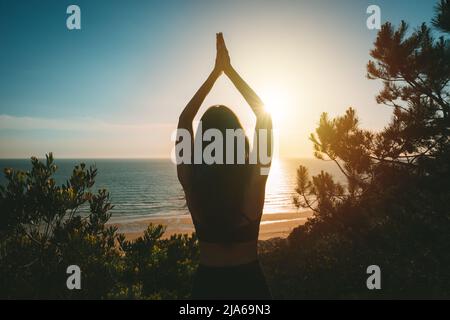 Silhouette of young healthy woman practicing yoga with ocean view at sunset. Rear view of woman standing with hands joined over head in tree pose Stock Photo