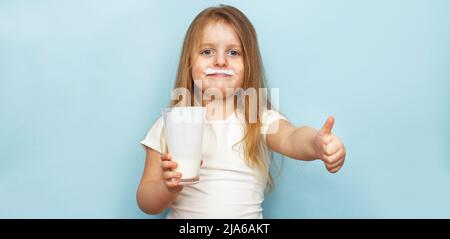 little beautiful girl holding milk in a glass on a blue background. child drinks yogurt. banner Stock Photo