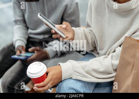 young people with smartphones and takeaway coffee sitting on a bench . Stock Photo
