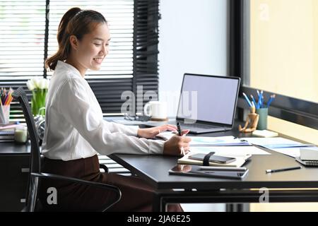 Side view female accountant using laptop computer and writing important information on notebook Stock Photo