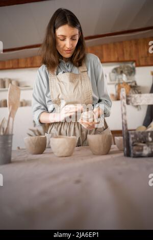 female artisan concentrated on modeling jug from raw clay while pottery lesson or workshop in creative studio. Woman ceramic business owner making Stock Photo