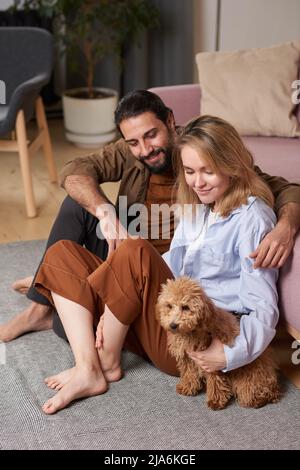 Young man and woman spending free time together at home sitting on floor in living room relaxing with their dog Stock Photo