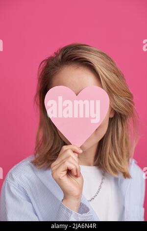 Medium close-up portrait of young Caucasian woman wearing casual outfit covering her face with paper heart Stock Photo