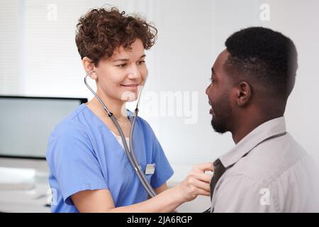 Horizontal medium close-up portrait of modern female doctor standing in front of male patient examining his heart beat and lungs using stethoscope Stock Photo