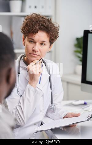 Vertical over-the-shoulder shot of serious therapist wearing white coat sitting in front of patient listening to patients health complaints and making Stock Photo
