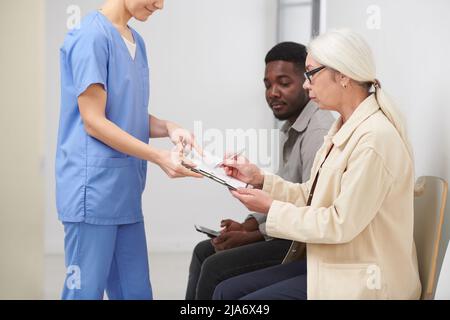Side view shot of stylish senior woman sitting in queue in hospital signing important medical document papers Stock Photo