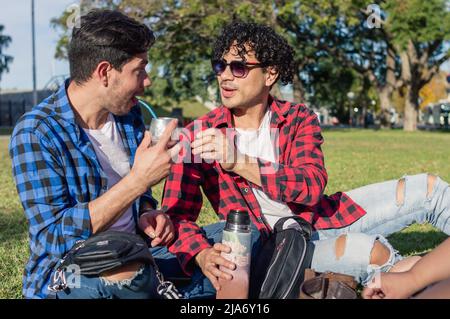 latin caucasian male homosexual argentinian couple sitting on the grass in the park smiling, enjoying drinking mate on a sunny day. Stock Photo