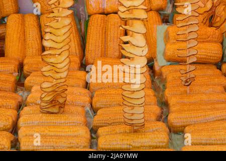 yellow cooked organic corn cobs with plastic wrap and some long skewers of fried potato chips with peel for sale in street food stall Stock Photo