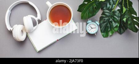 Workplace with a notepad, headphones, a cup of tea or coffee, alarm clock and green plant monstera. Freelancer working space on grey background. Top v Stock Photo