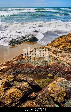 Detail of rock with puddle of water on sandy beach with ocean waves crashing Stock Photo