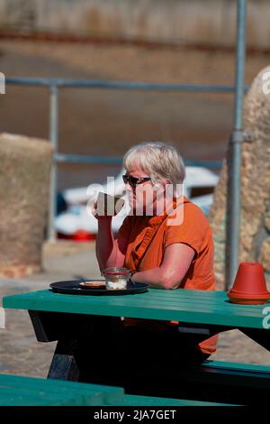 Elderly woman wearing an orange top and wearing sunglasses drinks a beverage on the harbour side in Porthleven, Cornwall Stock Photo