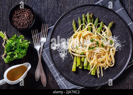 roasted asparagus spears with spaghetti sprinkled with shredded pecorino cheese and parsley on black plate on dark wooden table, horizontal view from Stock Photo