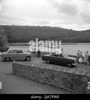 1950s, historical, people and cars of the era, including an Austin Cambridge, waiting at Feock, watching the King Harry Ferry, crossing the Carrick Roads reach of the estuary of the River Fal in Cornwall, England, UK. A small chain ferry bridge was established on this part of the estuary in 1888 using a steam engine to pull it along the chains and this was used until the introduction of a disel engine in 1956. One of the world's most scenic ferry trips, it is now one of only five chain ferries in England. Stock Photo