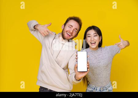 Asian girl and caucasian guy showing thumbs up holding smartphone showing blank white screen, mobile app advertisement and excited smile on camera isolated on yellow background. Product placement. Stock Photo