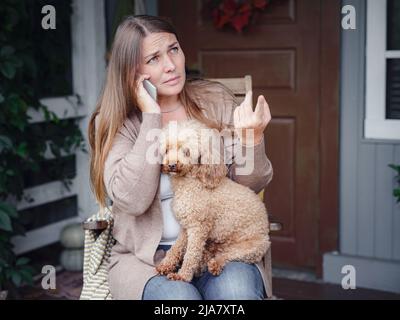 middle aged caucasian woman in brown cardigan sits on terrace of house with her beige poodle and emotionally speaks with friend via smartphone. morning good and bad news, autumn lifestyle concept Stock Photo