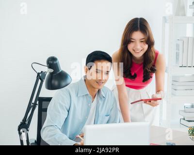 Happy smiling Asian business couple, young man and woman, colleagues using tablet and laptop computer on table, looking at screen, working together at Stock Photo