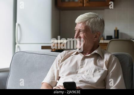 Smiling older age male pensioner sit on sofa hold smartphone Stock Photo