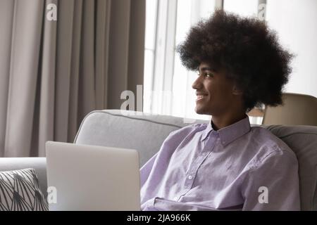 Happy dreamy Black laptop user sitting on couch, looking away Stock Photo
