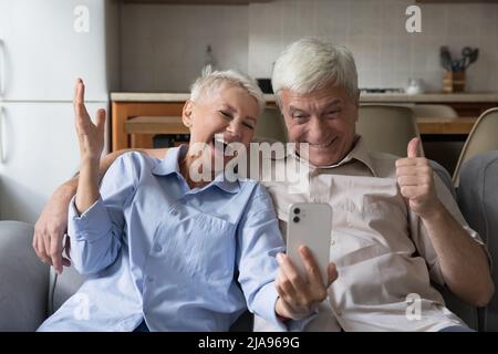 Overjoyed senior couple hug on sofa hold phone celebrate triumph Stock Photo