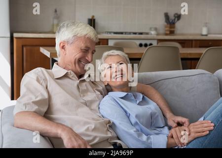 Laughing elderly spouses cuddle on couch at studio flat talk Stock Photo