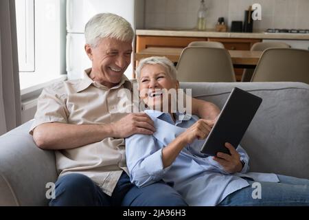 Excited older grandparents hug on couch laugh using digital touchpad Stock Photo