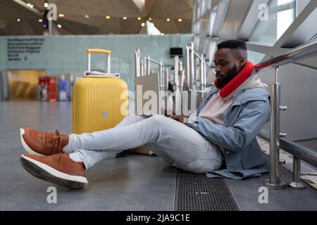African American hipster guy working at computer on knees sitting on floor near window in airport. Stock Photo