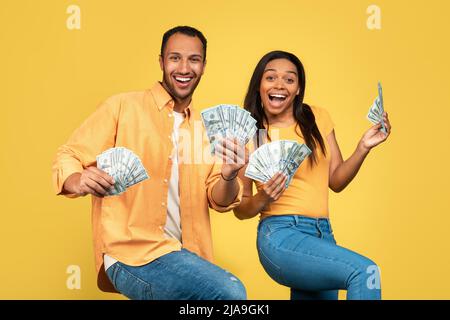 Excited young African American couple holding money, dancing and celebrating big win on yellow studio background Stock Photo