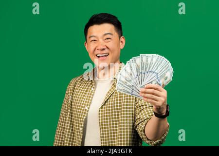 Happy middle aged asian man holding fan of money cash in hand and smiling at camera, posing on green background Stock Photo
