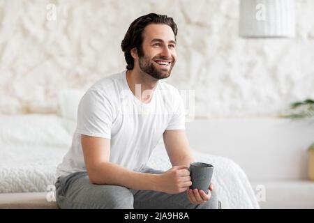 Happy bearded young man sitting on bed, drinking coffee Stock Photo