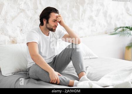 Exhausted young man sitting on bed, touching his head Stock Photo