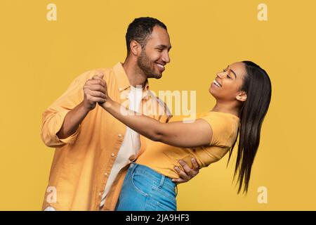 Positive young African American couple in casual outfits dancing and smiling at each other on yellow background Stock Photo