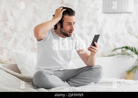 Shocked young man sitting on bed with smartphone, touching head Stock Photo