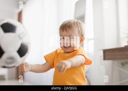 Joyful little boy playing with football ball, having fun and enjoying game at home, living room interior Stock Photo