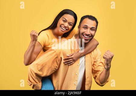 Attractive young African American man giving piggyback ride to his girlfriend, gesturing YES on yellow studio background Stock Photo