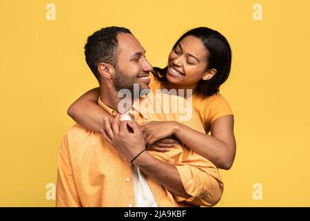 Affectionate young black woman hugging her boyfriend, looking in his eyes on yellow studio background Stock Photo