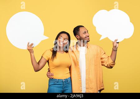 Happy young black couple holding empty speech bubbles on yellow studio background, mockup Stock Photo