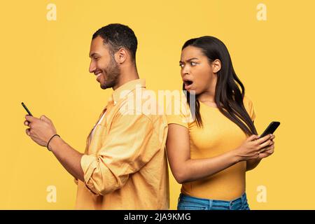Shocked black woman spying on her boyfriend who is using smartphone, chatting with lover, cheating on yellow background Stock Photo