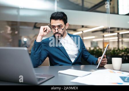 Serious busy attractive millennial arab businessman with beard in glasses works with computer and documents Stock Photo